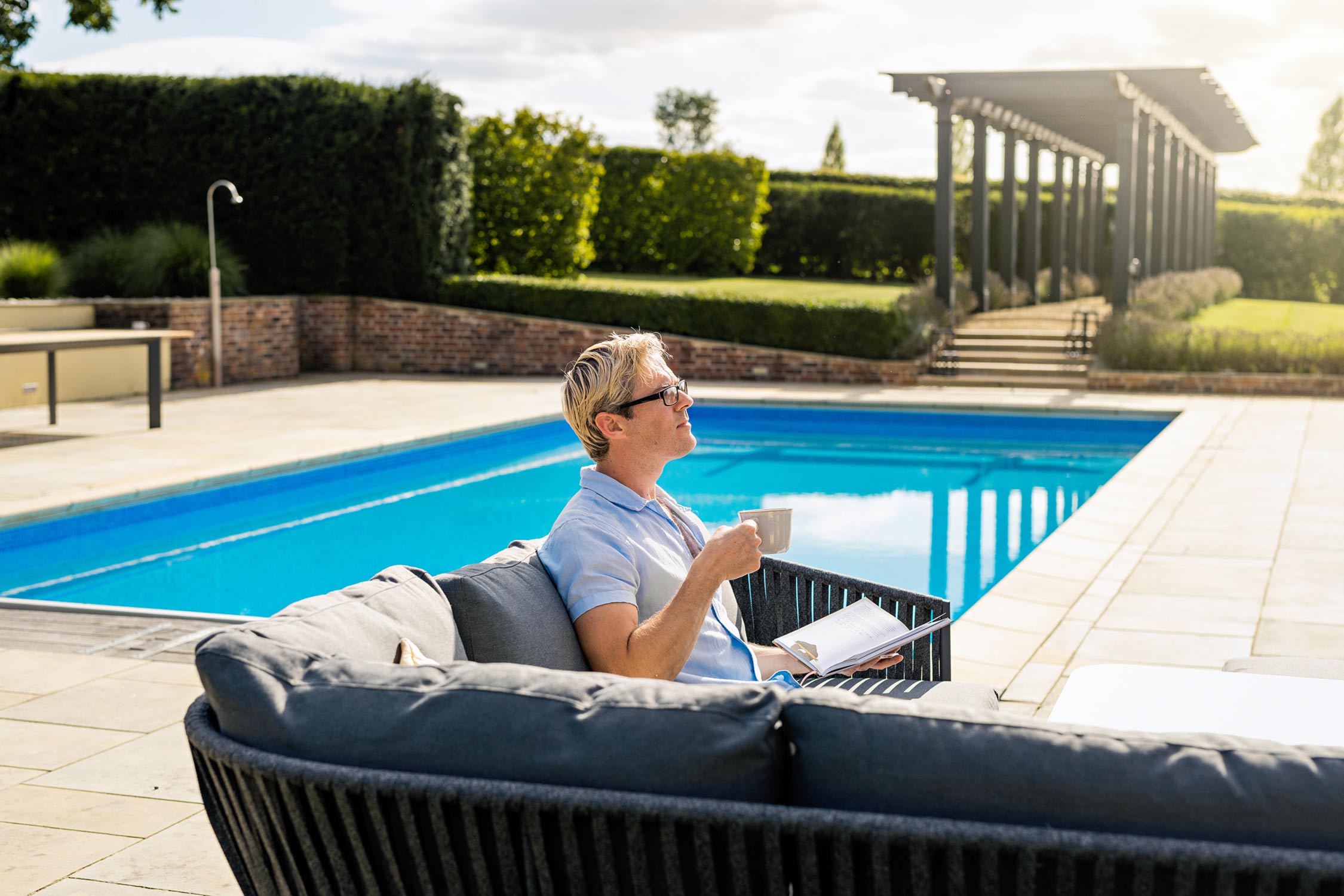 Lifestyle product photography of a high end garden furniture with an attractive man drinking coffee by a swimming pool in a smart summer garden.