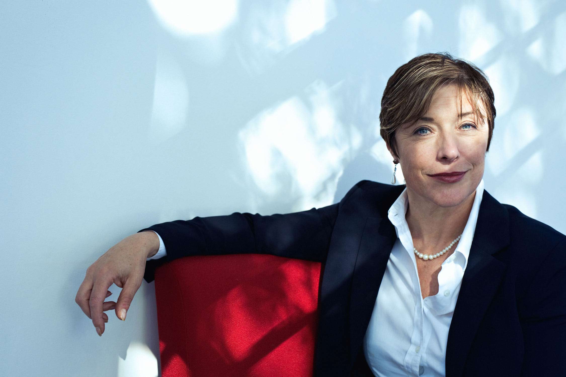 Moody modern corporate portrait of a female CEO sitting on a red seat with light and shade hitting her face and the wall behind.