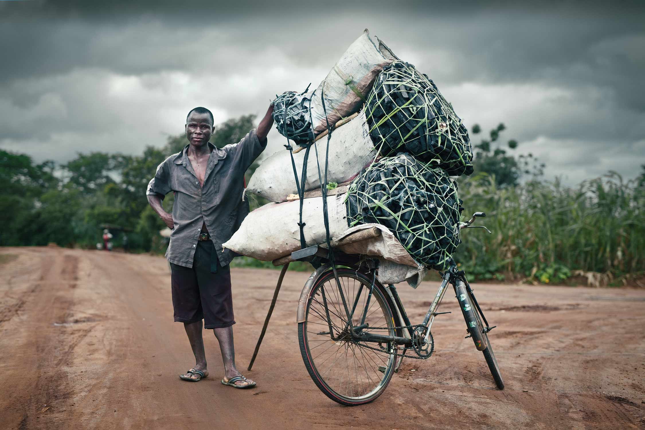 Stylised documentary portrait of rural Malawian charcoal seller standing by his charcoal-laden bicyle.