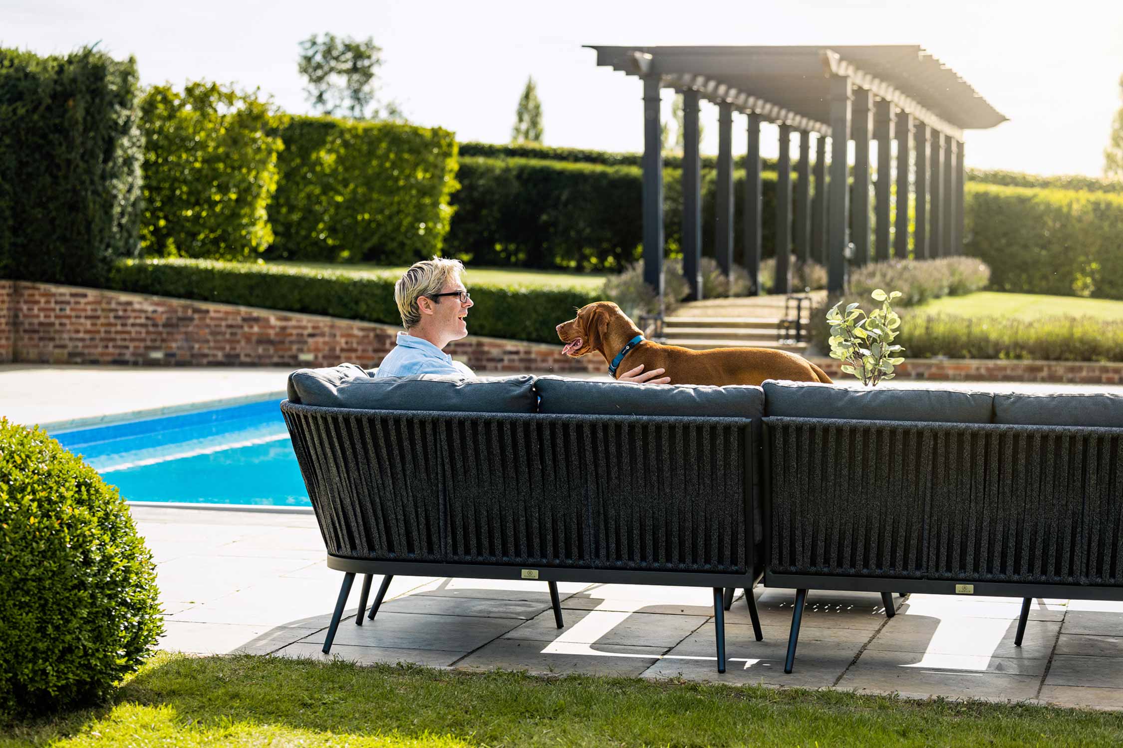 Lifestyle product photography of a high end garden furniture with a man and dog by a swimming pool in a smart summer garden.
